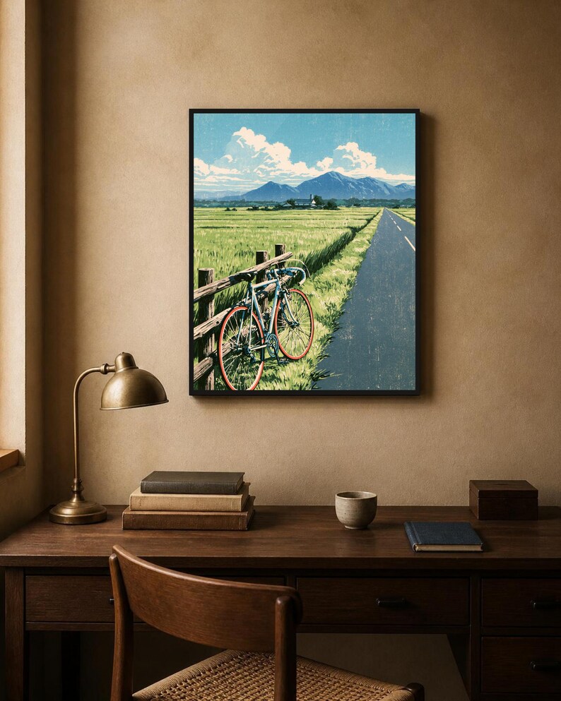 May include: A framed wall art print depicting a light blue bicycle with red wheels leaning against a wooden fence beside a paved road. The scene features a vast green field, distant mountains, and a blue sky with white clouds in a landscape orientation.