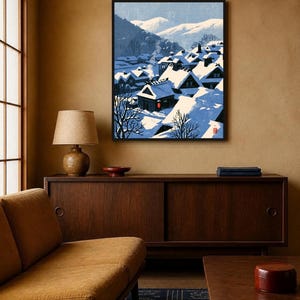 May include: A framed wall art print depicting a snowy Japanese village scene with traditional gabled houses covered in deep snow, blue shadows, and distant mountains under a pale sky, featuring a small red circular seal in the bottom right corner.