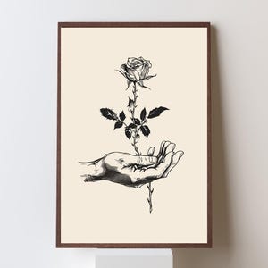 May include: A framed wall art print featuring a black and white ink illustration of a hand holding a single rose with thorns. The artwork is set against a solid cream-colored background, enclosed within a dark brown wooden frame.