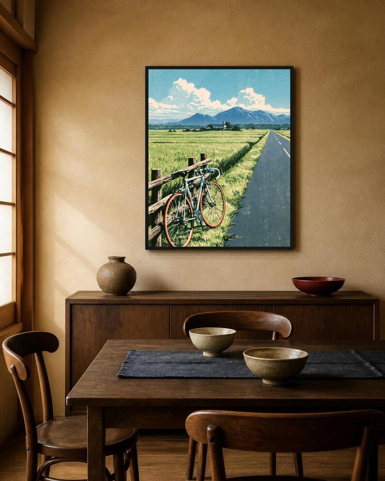 May include: A framed wall art print depicting a landscape with a road, a green field, and a bicycle leaning against a wooden fence. The scene features distant blue mountains under a bright blue sky with white clouds, rendered in a graphic style.