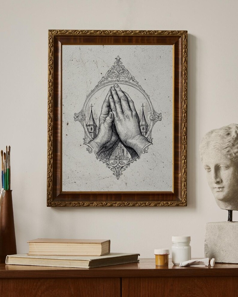 Op de afbeelding: Een ingelijste wandkunstprint met een zwart-witte inktillustratie van twee handen die in gebed zijn samengevouwen. De handen zijn gecentreerd in een sierlijke, ovale lijst, met silhouetten van kerktorens zichtbaar op de achtergrond.