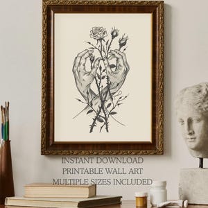 May include: A framed wall art print featuring a black and white line drawing of two hands gently holding a rose stem with thorns and buds. The artwork is displayed in a dark brown ornate wooden frame with a cream-coloured background.