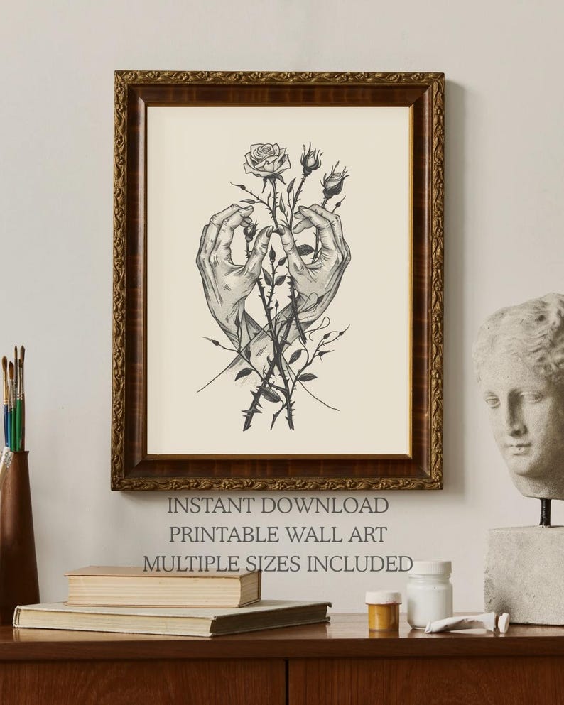 May include: A framed wall art print featuring a black and white line drawing of two hands gently holding a rose stem with thorns and buds. The artwork is displayed in a dark brown ornate wooden frame with a cream-coloured background.