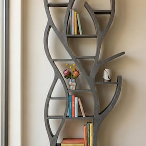 May include: A grey wooden bookcase with an unusual tree-shaped design. The shelves display colourful books and a small vase with flowers. The bookcase is positioned near a window, enhancing the natural feel of the room.