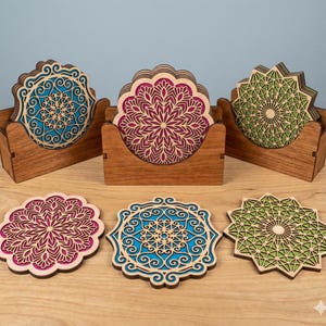 May include: Laser-cut wooden coasters featuring mandala patterns in teal, magenta, and green. The coasters are displayed in wooden holders. The coasters are on a light wood surface, showcasing their decorative appeal. Each coaster has a unique, detailed design.
