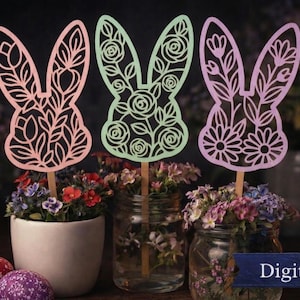 May include: Three decorative Easter bunny cutouts in pink, green, and lavender, each with floral designs. The bunnies are on wooden sticks, placed in flower arrangements. The words "Digital File" are in the bottom right corner.