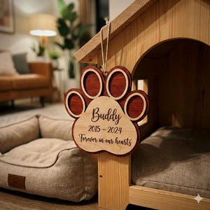 May include: A wooden paw print-shaped memorial sign with the name "Buddy" and dates 2015-2024, hanging from a dog house. The sign reads "Forever in our hearts." The sign is made of light and dark brown wood.