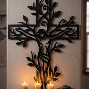 Könnte beinhalten: Schwarzes Metall-Wandkreuz mit einem Lebensbaum-Design. Das Kreuz ist an einer beigefarbenen Wand befestigt, mit drei brennenden Kerzen und einem Buch auf einem Holzregal darunter. Das Kreuz hat filigrane Ausschnitte von Blättern und Ästen.