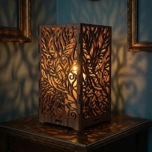 May include: A wooden lamp with a rectangular shape and intricate leaf-like cutouts. The lamp emits a warm, inviting glow. It sits on a dark wooden table, creating a cozy ambiance. The lamp's design suggests a nature-inspired theme.