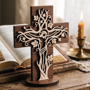 May include: A wooden cross with a tree of life design, featuring a cutout of a figure. The cross is brown with a lighter wood design and stands on a round base. A lit candle and an open book are in the background.
