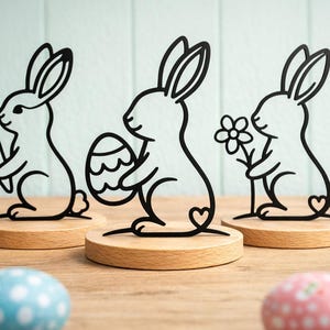 May include: Three black metal Easter bunny figurines on wooden bases. Each bunny holds a different item: a carrot, an Easter egg, and a flower. Pastel-colored Easter eggs are in the foreground. The bunnies are approximately 10 cm tall.