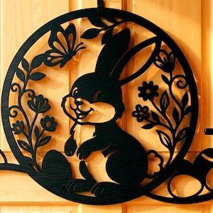 Peut inclure: Décoration de porte en métal noir représentant un lapin de Pâques. Le design circulaire présente un lapin souriant, un papillon, des fleurs et des œufs de Pâques. La découpe est fixée sur une porte en bois.