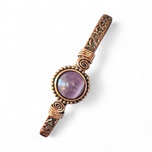 May include: A copper-colored hair barrette with an oval, polished, purple gemstone in the center. The barrette has decorative spiral wire accents and etched patterns on the ends.