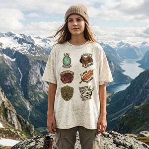 Op de afbeelding: Een off-white t-shirt met korte mouwen en een verweerde, gespikkelde textuur. De voorkant bevat zes grafische patches met de tekst: Egg Bound, Rider's Bond, Skyborne, Flame Bound, Scale Warden en Dragon Rider Ride or Die.