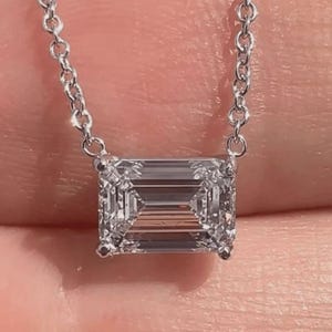 May include: A delicate silver necklace featuring a rectangular, emerald-cut diamond pendant. The diamond is clear and faceted, reflecting light. The chain is fine and links together. The necklace is displayed against a soft, blurred background.