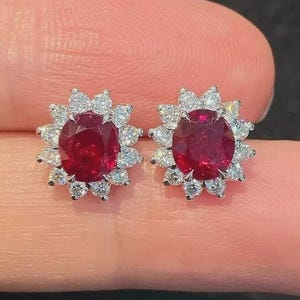 Può includere: Un paio di orecchini a lobo con pietre preziose rosse ovali, ciascuna circondata da un alone di pietre preziose chiare più piccole. Gli orecchini sono incastonati in un metallo color argento, creando un design scintillante ed elegante.