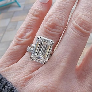 May include: A large emerald-cut diamond ring featuring a prominent rectangular center stone set in a silver-toned metal band. The ring is accompanied by a secondary band adorned with smaller round diamonds, creating a layered appearance.