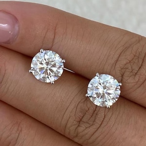 May include: A pair of round, clear diamond stud earrings. The earrings are set in a silver metal. The diamonds sparkle and reflect light. The earrings are displayed on a hand with a light pink manicure.