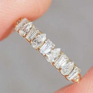 May include: A gold band ring set with white diamonds of different shapes. The ring is held between two fingers with light blue nail polish. The brand name "ROSSY JewelsIN" is in the upper right corner.