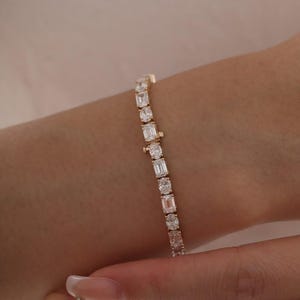 May include: A gold bracelet featuring a series of alternating round and emerald-cut clear gemstones. The bracelet is displayed on a wrist, showcasing the sparkle of the stones.