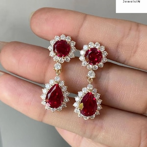 Pode incluir: Um par de brincos pendurados dourados com grandes pedras preciosas vermelhas em forma de gota, rodeadas por um halo de pequenas pedras brancas brilhantes. Design floral no topo e em forma de gota na parte inferior.