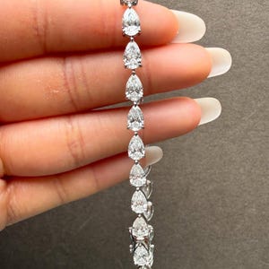 May include: A silver-toned earring featuring a series of pear-shaped, clear gemstones. The gemstones are arranged in a vertical line, with a small, square-cut gemstone at the top. The earring is held against a neutral background.