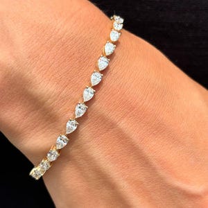 Puede incluir: Un brazalete dorado con una serie de piedras preciosas claras en forma de pera. Las gemas están engastadas juntas, creando una línea continua de brillo. El brazalete está en una muñeca, sobre un fondo negro.