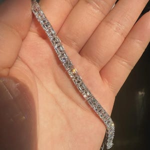 May include: A dazzling silver bracelet featuring multiple square-cut, clear gemstones. The bracelet is held in a hand, highlighting its reflective properties. The gemstones are set closely together, forming a continuous line of sparkle.