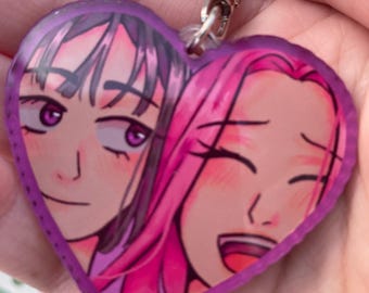 May include: Heart-shaped keychain featuring a colorful illustration of two anime-style women. One woman has purple hair and the other has pink hair. The keychain has a silver clasp.