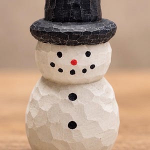 May include: A small, hand-carved wooden snowman figurine with a textured, off-white finish. The snowman wears a black top hat and features a painted face with black dot eyes, a small red nose, and a black dot smile, plus three black buttons.