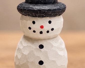 May include: A small, hand-carved wooden snowman figurine with a textured, off-white finish. The snowman wears a black top hat and features a painted face with black dot eyes, a small red nose, and a black dot smile, plus three black buttons.