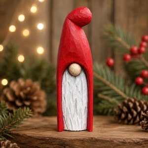 May include: A hand-carved wooden gnome figurine featuring a tall, smooth red hat that curves downward at the top. The gnome has a small, round, natural wood-toned nose and a long, textured white beard carved into the front of the red body.