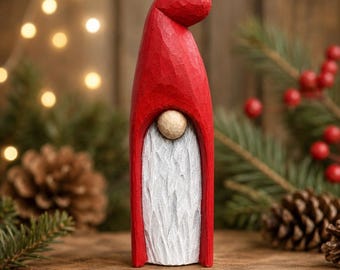 May include: A hand-carved wooden gnome figurine featuring a tall, smooth red hat that curves downward at the top. The gnome has a small, round, natural wood-toned nose and a long, textured white beard carved into the front of the red body.