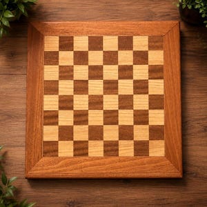 May include: A square wooden chessboard featuring a classic alternating grid pattern of light tan and medium brown squares. The board is framed by a wide, solid border made of polished wood with a visible grain, creating a clean, flat surface.