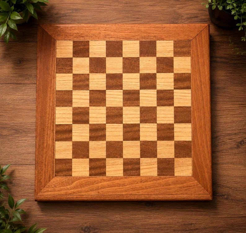 May include: A square wooden chessboard featuring a classic alternating grid pattern of light tan and medium brown squares. The board is framed by a wide, solid border made of polished wood with a visible grain, creating a clean, flat surface.
