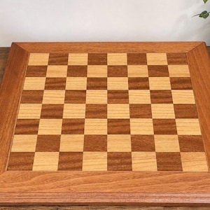 May include: A square wooden chessboard featuring a classic alternating grid pattern of light tan and medium brown wood squares. The board is framed by a wide, smooth border made of the same medium brown wood, creating a clean and uniform finish.