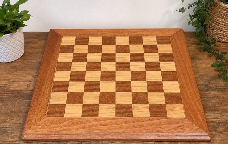 May include: A square wooden chessboard featuring a classic alternating grid pattern of light tan and medium brown wood squares. The board is framed by a wide, smooth border made of the same medium brown wood, creating a clean and uniform finish.