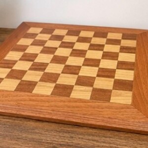 May include: A square wooden chessboard featuring a classic alternating grid pattern of light tan and medium brown wood squares. The board is framed by a wide, smooth border made of a single tone of medium brown wood with visible grain.