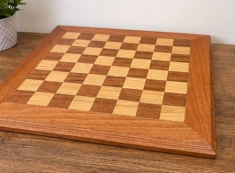 May include: A square wooden chessboard featuring a classic alternating grid pattern of light tan and medium brown wood squares. The board is framed by a wide, smooth border made of a single tone of medium brown wood with visible grain.