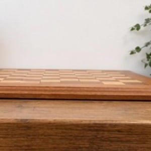 May include: A rectangular wooden chessboard featuring a classic alternating grid pattern of light and dark brown squares. The board has a smooth, polished finish with a thick, beveled wooden frame surrounding the playing surface area.