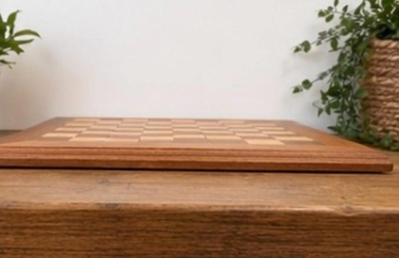 May include: A rectangular wooden chessboard featuring a classic alternating grid pattern of light and dark brown squares. The board has a smooth, polished finish with a thick, beveled wooden frame surrounding the playing surface area.