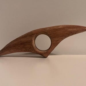 May include: A wooden book page holder with a natural finish. The holder has a curved, wing-like shape with a circular opening for the thumb. The wood grain is visible, adding texture to the design.