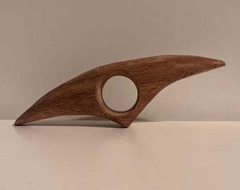 May include: A wooden book page holder with a natural finish. The holder has a curved, wing-like shape with a circular opening for the thumb. The wood grain is visible, adding texture to the design.