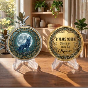 May include: Two circular medallions on clear stands. The left medallion features a blue wolf howling at a full moon within a gold and teal ornate border. The right medallion displays the text 2 YEARS SOBER, Choose joy every day, and Melissa.