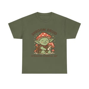 Könnte beinhalten: Olivgrünes T-Shirt mit einer Cartoon-Figur mit Pilzkopf, die einen Pilz hält. Der Text "SHROOM DRUID" und "SPREADING SPORES & JOY" ist über und unter der Figur aufgedruckt.