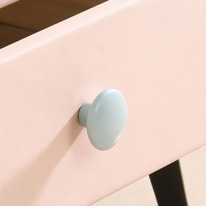 May include: Close-up of a pale pink chest of drawers with two drawers. Each drawer features a light blue, oval knob. One drawer is open, showing the wooden interior. The chest has black legs.