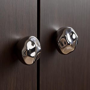 May include: Two polished chrome cabinet knobs with an organic, textured design are mounted on a dark brown wooden cabinet door. The knobs reflect light, creating a contrast in colour and texture. The cabinet door is made of wood.