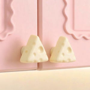 May include: Two off-white, cheese-shaped cabinet knobs with holes are mounted on a light pink cabinet. The cabinet doors have a scalloped edge design. The knobs are a whimsical touch for kitchen or children's furniture.