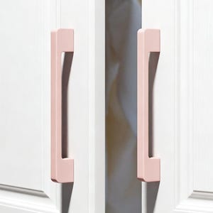 Puede incluir: Dos tiradores de armario de color rosa pálido están montados en una puerta de armario blanca. Los tiradores son rectangulares con un asa cuadrada. La puerta del armario está ligeramente entreabierta, revelando un vistazo del interior.