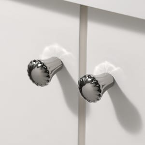 以下が含まれることがあります： 溝のあるデザインの、2 つの銀色のキャビネットノブのクローズアップ。ノブは白いキャビネットドアに取り付けられており、それらの間に垂直のサポートビームが見えます。ノブは反射面を持っています。